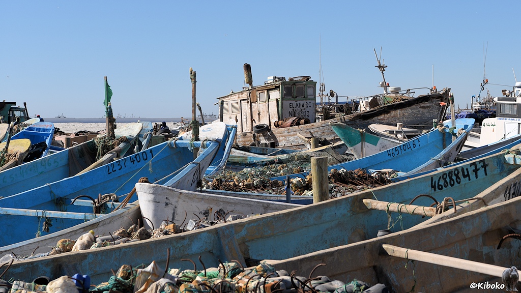 Das Bild zeigt einige offene Fischerboote mit hellblau gestrichenen Innenseiten. In den Booten liegen bunte Plastikschiwimmer und rostige Metallhaken. Im Hintergrund ist ein etwas größeres, hölzernes Fischerboot mit der Aufschrift Elkhair 1.