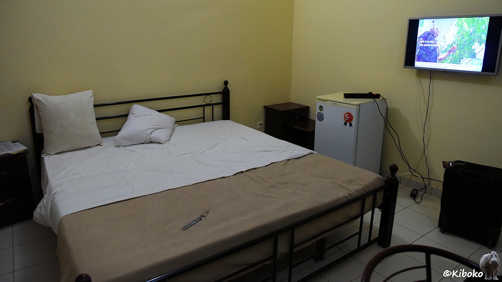Das Bild zeigt ein Hotelzhimmer mit Doppelbett im Metallgestell mit hellbrauner Decke, einen Kühlschrank und einen an der Wand montierten Flachbildschirm.