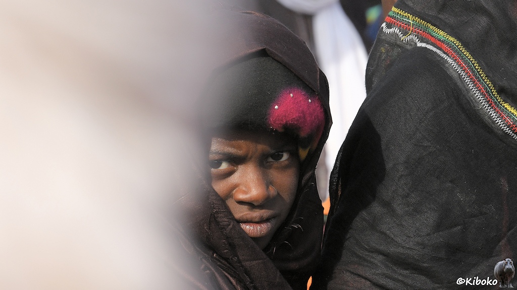 Das Bild zeigt das Porträt einer jungen Frau im dunklen Gewand. Ein schwarzes Tuch vor der Stirn hat einen großen rosa Fleck. Der Gesichtsausdruck ist sehr nachdenklich. Im linken Bildbereich ist ein heller, unscharfer Bereich einer vorbeigehenden Person.