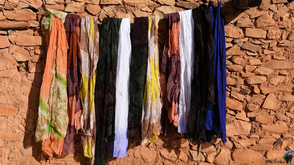 Das Bild zeigt aufgehängte Tücher vor einer rötlichen Natursteinwand. Die Tücher sind schwarz, weiß, blau, gelb-braun gemustert und orange-grün gemustert.