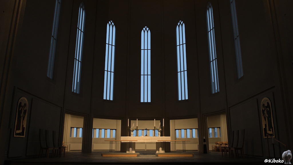 Der Altarraum ist dunkel. Der schlichte Altar ist hell erleuchtet. Dahinter sind 7 langgestreckte Fenster mit Klarglas.