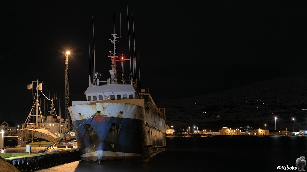 Ein blau-weißer Fischtrawler liegt an der Hafenmole. Im Hintergrund ist ein weiteres kleines Schiff und Häuser mit Weihnachtsdekoration.