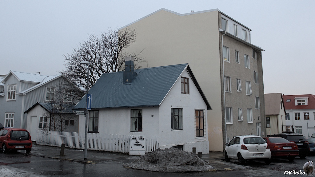 Ein kleines weißes Holzhaus mit schwarzen Fensterrahmen und blaugrauem Blechdach steht an einer Sraßenkreuzung. Das kleine Grundstück ist mit einem weißen Lattenzaun umgeben. Dahinter sind ein graues Holzhaus und ein beigefarbener Wohnblock. Davor stehen Autos und liegt ein Haufen dreckiger Schnee.