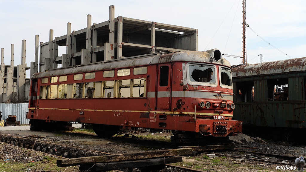 Das Bild zeigt den ausgeschlachteten Kasten einer roten Elektrolokomotive vor einer Bauruhine mit in Himmel wachsenden Betonstützen.