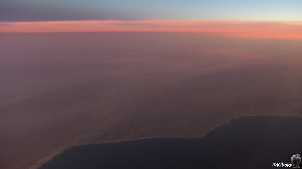 Das Bild zeigt eine Luftaufnahme einer braunen Wüstenlandschaft. Im Vordergrund ist das Meer. Ein Wolkenschleier über der Wüste leuchtet lachsfarben.