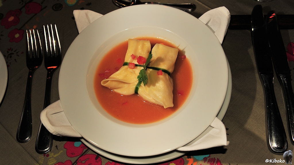 Das Bild zeigt einen Teller mit einem großen zusammengefalteten, quadratischen Teigtasche, die mit einen Schnittlauchstreifen zusammengehalten wird. Die Teigtasche schwimmt in einer rotorangen Suppe.