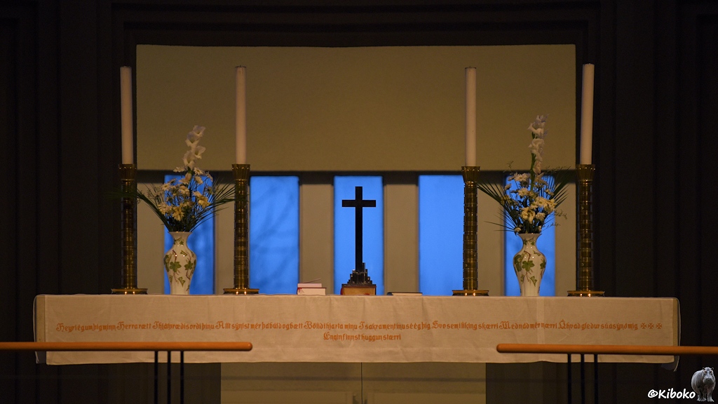 Auf einem schlichten Altar stehen ein Kreuz, zwei Vasen mit weißen Blumen, sowie vier hohe Kerzenständer mit weißen Kerzen.