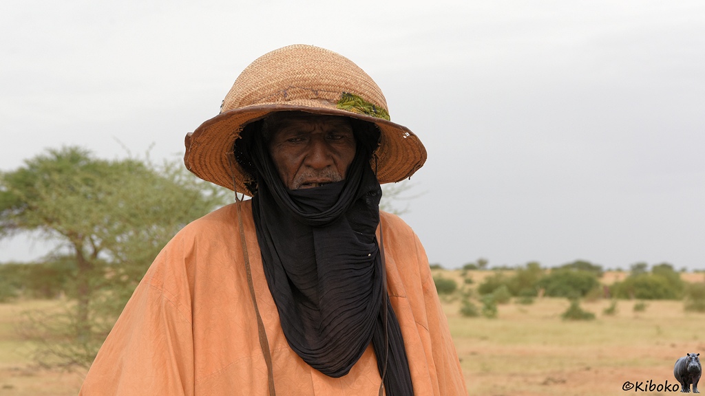 Das Bild zeigt das Porträt eines älteren Mannes mit orangenem Gewandt, schwarzen Turban der auch um den Hald und über dem Mund gewickelt ist. Dazu trägt er einen geflochtenen Stohhut in der Form einer Melone.