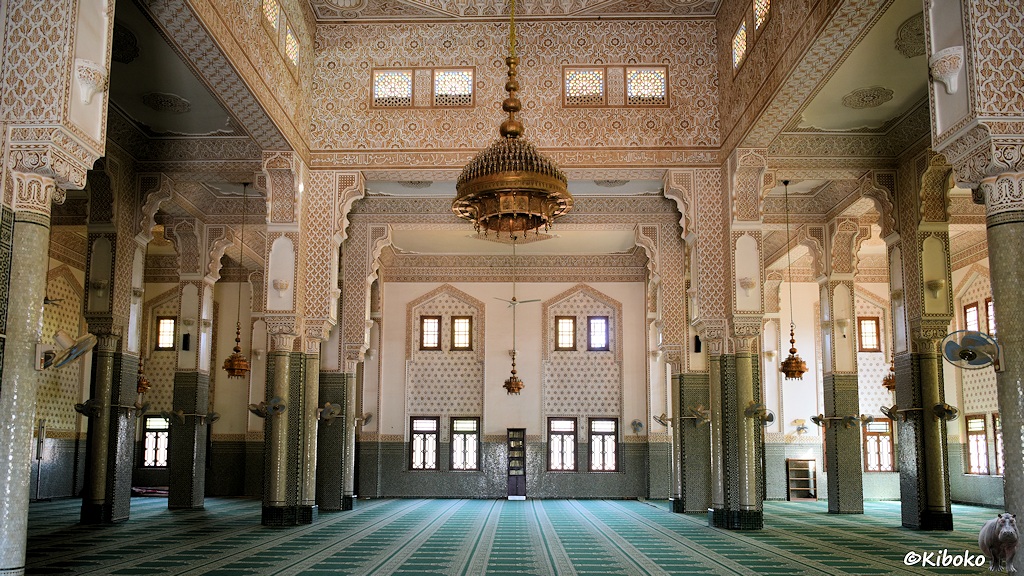Das Bild zeigt die Innenansicht der großen Moschee. Die rechteckige Halle wird durch zwei Reihen Säulen, die mit hellgrünen Mosaiken verziert sind unterteilt. Der Boden ist mit einem grünen Teppich belegt, der mit hellen Streifen die Plätze für die Gläubigen aufteilt. Die Wände und Decken sind mit hellbraunen Mosaiken bedeckt. In der Mitte hängt ein großer, messingfarbener Kronenleuchter.