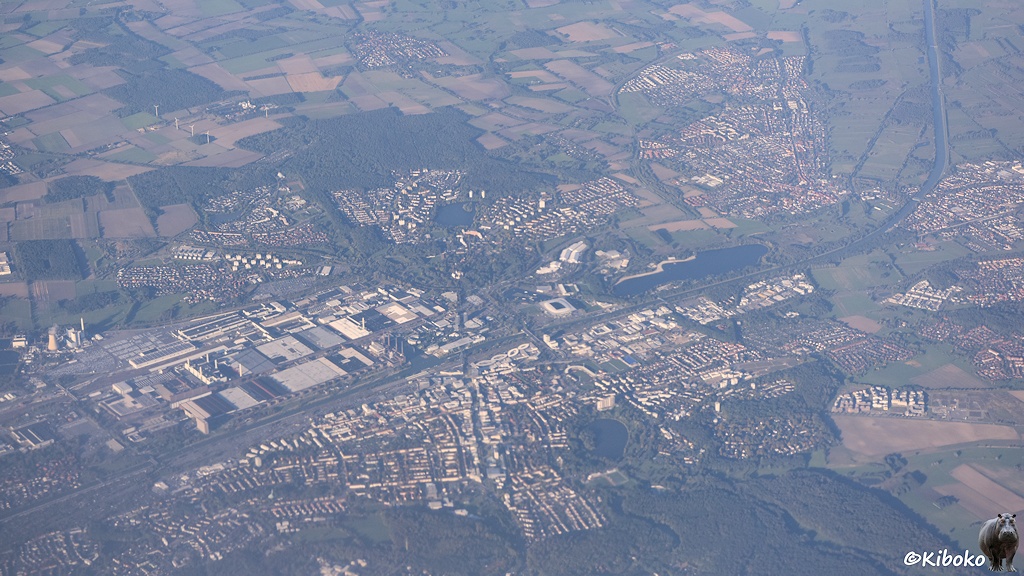 Das Bild zeigt eine Luftaufnahme aus großer Höhe von einer Stadt, die durch einen Kanal geteilt ist. Auf der anderen Kanalseite ist eine große Fabrik.