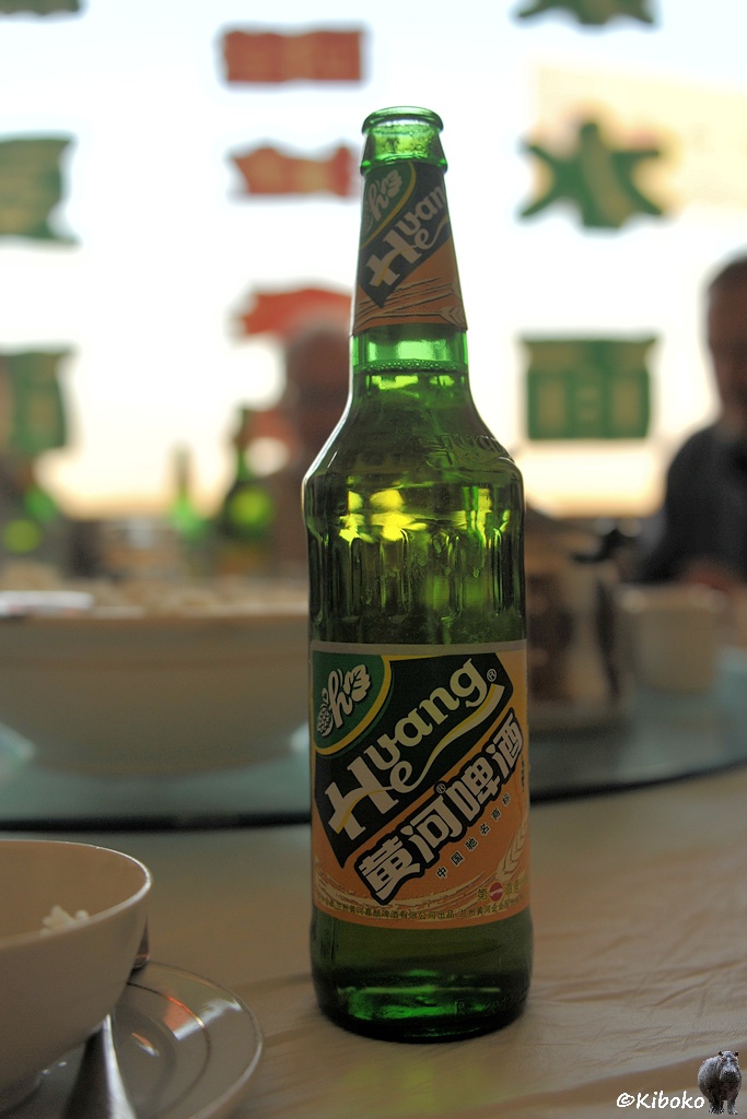 Das Bild zeigt eine grüne Bierflasche mit orangenem Ettikett. Darauf sind diagonal schwarze Felder mit weißer Schrift: Huang He. Im Hintergrund sind unscharf chinesische Schriftzeichen an einer Glasscheibe.