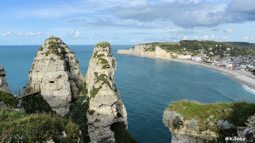 Das Bild zeigt eine Strandbucht mit einem Ort am rechten Bildrand. Im Hintergrund ist eine steilküste aus hellem Sandstein. Im Vordergrund sind links zwei Felsnadeln und rechts eine grawbeachsener Fels.