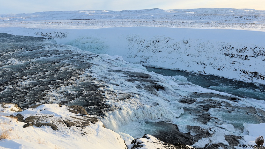 Das Bild zeigt einen Wasserfall von oben. Das Wasser fließt über Stromschnellen und kleine Stufen im Vordergrund. Dahinter stürzt es in eine 11m tiefe Schlucht, die nicht einsehbar ist.