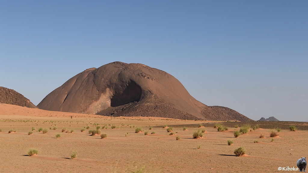 Das Bild zeigt einen kahlen, rotbraunen Berg in einer hellbraunen Sandwüste mit einzelnen Grasbüscheln.