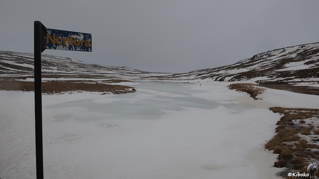 Ein mit Schnee und Eis bedeckter Fluss windet sich zwischen sanften mit Schneeresten bedeckten Hügeln. Im Vordergrund ist ein Schild mit der Aufschrift Norðurá.