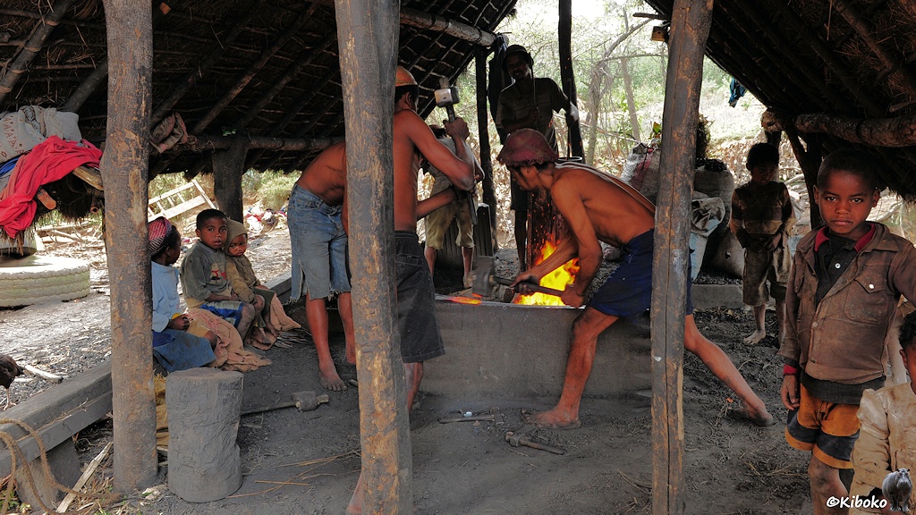 Das Bild zeigt eine Feuerstelle in einer offenenm aufgeständerten Hütte. Drei Männer bearbeiten ein glühndes Metallstück vor einem Feuer. Im Hintergrudn betätigt ein Mann eine manuelle Luftpumpe. Kinder sitzen auf beiden Seiten und beobachten die Männer.