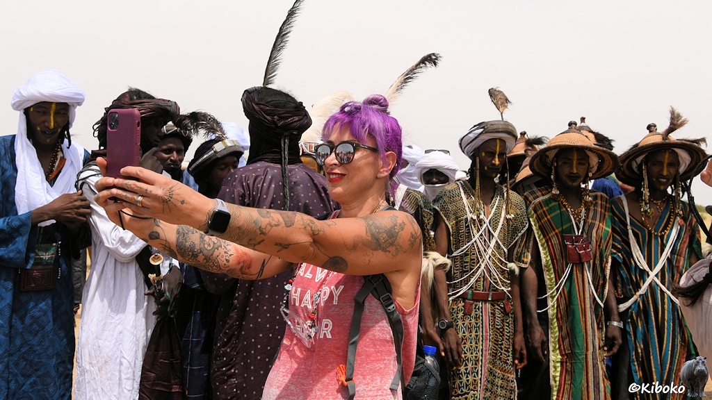 Das Bild zeigt eine tätowierte Touristin mit lila gefärbten Haaren und Spiegelbrille. Sie hält ein Handy mit ausgestreckten Armen vor sich. Im Hintergrund stehen drei Männer im traditionellen Gewandt und schauen etwas skeptisch der Touristin zu. Weitere Männer haben ihr den Rücken zugedreht.