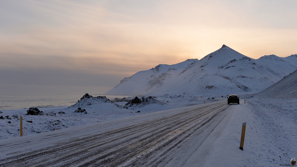 Das Bild zeigt ein parkenden Kleinwagen an einer vereisten Straße im Gegenlicht. Links ist das Meer. Rechts ist eine Bergkette, hinter der sich die Sonne befindet.