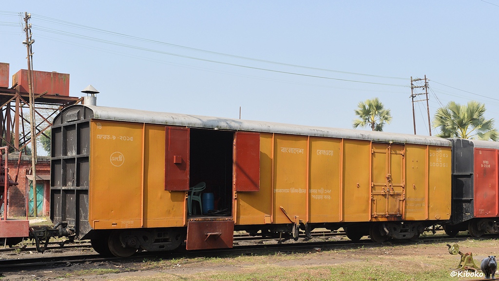 Das Bild zeigt einen gelben vierachsigen gedeckten Güterwagen. Der Wagen hat schwarze Enden und ein silbernes Dach. Die linke Tür steht offen. Beide Flügel sind aufgeklappt und der Unterteil ist nach untengeklappt. Im Wagen stehen Plastikstühle. Der Wagen dient als Gerätewagen zum Kranzug.