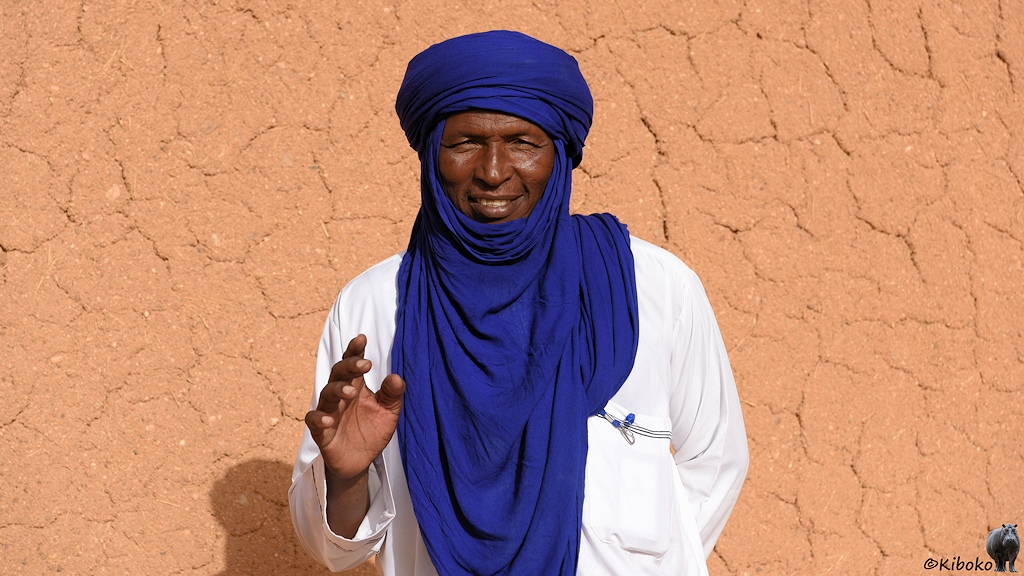 Das Bild zeigt einen älteren Mann im weißen Gewandt mit blauem Turban vor einer Lehmwand. Er lächelt und hebt die Hand zum Grüßen.