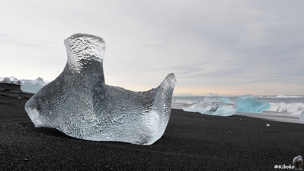 Das Bild zeigt einen kleineren Weißen Eisblock aus niedriger Perspektive am schwarzen Strand. Der Eisblock hat zwei Spitzen und eine Textur aus kleinen Dellen.