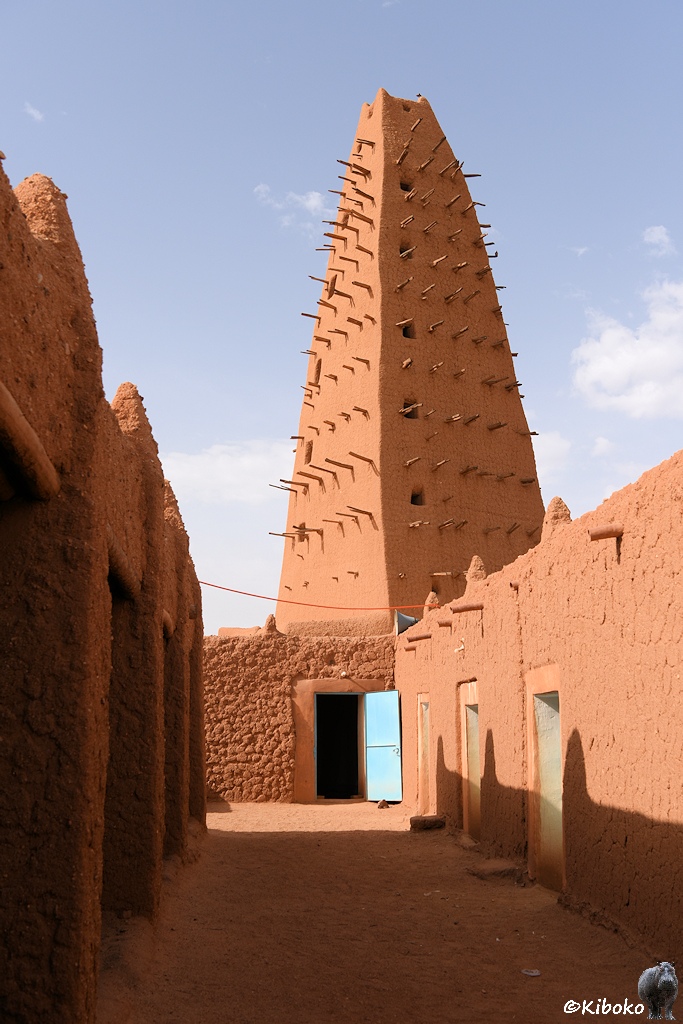 Das Bild zeigt eine Hochformataufnahme eines Zugangs zu einer Moschee mit einstöckigen Gebäuden aus rotbraunem Lehm auf beiden Seiten. Der Gang endet an einem einstöckigen Gebäude mit geöffneter hellblauer Tür. Darüber ragt ein viereckiger Turm aus rotbraunem Lehm auf. Der Turm verjüngt sich leicht nach oben. Auf jeder Seite ragen in waagerechten Reihen Knüppel aus der Wand. Sie haben einen Abstand von ungefäht einen halben Meter und sind bis zu 1 Meter lang. Der Turm hat 13 Reihen Knüppel.