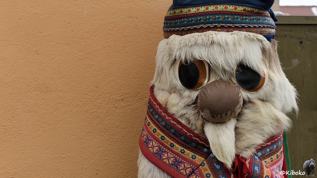 Das Bild zeigt das Porträt einer Trollfigur aus weißem Rentierfell. Sie trägt eine bunte Mütze und einen bunten Schal.