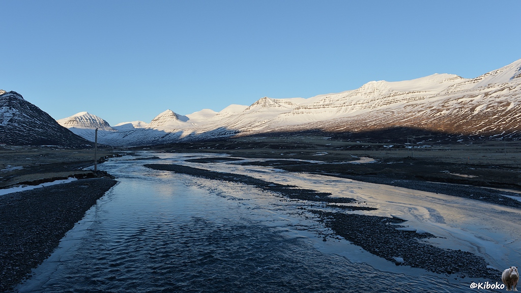 In einem schattigen Tal läuft ein flacher Fluss. Im Fluss sind Schotterinseln. Seitenarme des Flusses sind zugefroren. Die verschneiten Berge im Hintergrund werden von der Sonne angestrahlt.