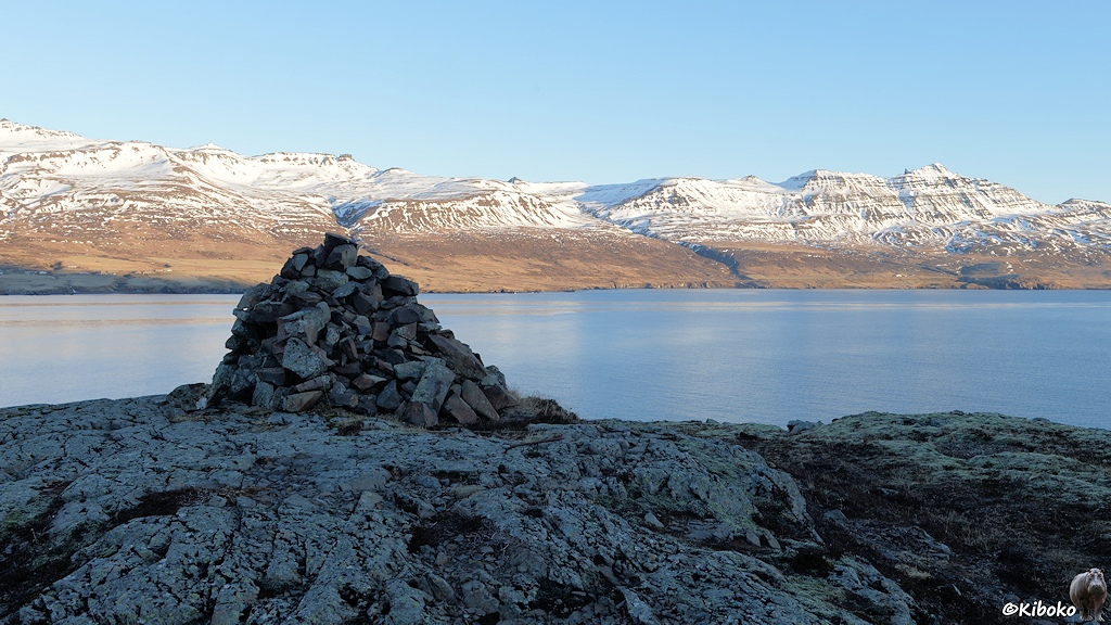 Auf einem Felsen ist ein großer Steinhaufen. Dahinter ist ein Fjord. Das gegenüberliegende Ufer liegt in der Sonne. Unten sind Wiesen. Oben sind schneebedeckte Berge.