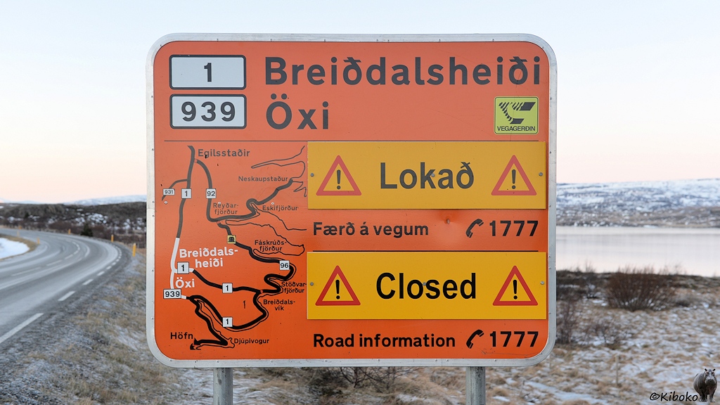 Ein großes Oranges Hinweisschild steht am Straßenrand. Es zeigt mehrere Alternative Straßenverläufe. Die kürzesten Strecken haben Aufsteckschilder mit der Aufschrift Lokað und Closed.