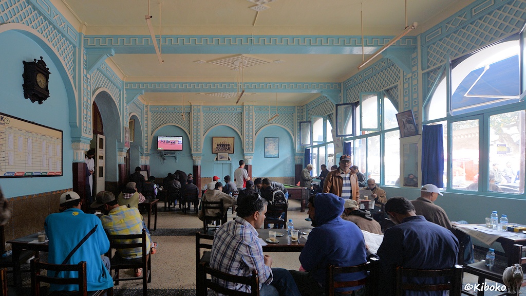 Das Bild zeigt die Innenansicht eines Cafés. Die Wände sind hellblau gestrichen und mit Bögen und Mustern weiß verziert. An kleinen Tischen sitzen Männergruppen bei Kaffee und Mineralwasser. Auf der Rückwand ist ein Flachbildfernseher mit Bilder vom Pferderennen.