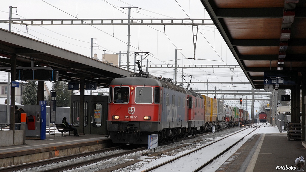 Das Bild zeigt eine rot-blaue und eine rote Elektrolokomotive vor einem Containerzug bei der Durchfahrt durch einen Bahnhof.