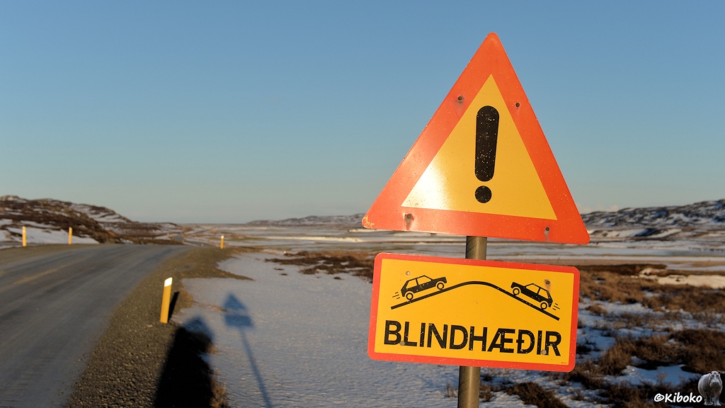 Die Straße führt über eine Kuppe. Am Straßenrand steht ein dreieckiges gelbes Schild mit rotem Rand und schwarzem Ausrufezeichen. Ein gelbes Zusatzschild mit rotem Rand zeigt zwei Autos die aufeinander zufahren. Dazwischen ist eine stilisierte Kuppe. Darunter ist die Aufschrift BLINDHÆÐIR.