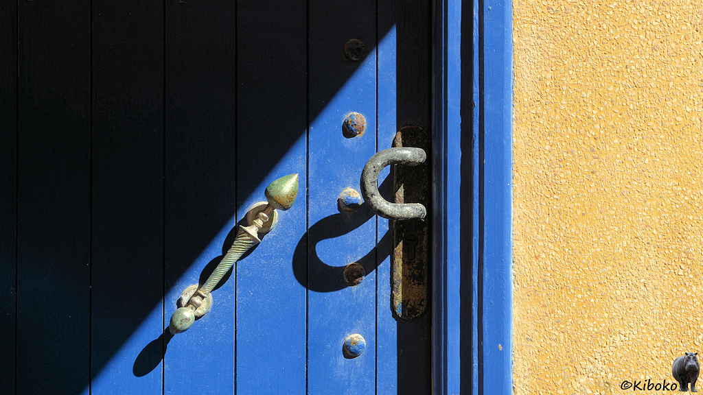 Das Bild zeigt zeigt einen Ausschnitt einer blauen Tür mit Schattenspiel. Der linksobere Teil ist im dunklen Schatten. Parallel zur Schattenkante ist ein Messingfarbener Türgriff in der Sonne. Am rechten Bildrand ist ein Streifen aus gelb gestrichenen groben Putz.