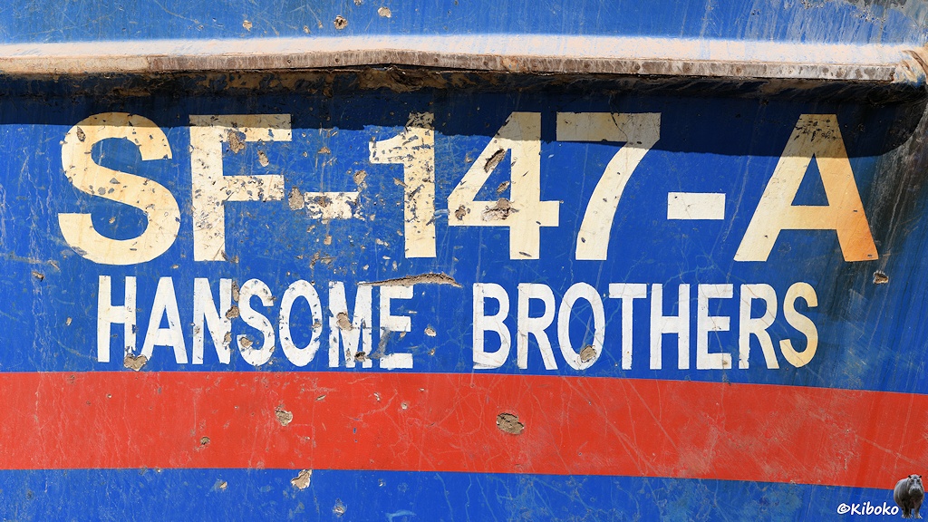 Das Bild zeigt die Anschrift an einen Bug eines Fischtrawlers. Auf blauem Grund steht in weißer Schrift SF-147-A und etwas kleiner darunter HANSOME BROTHERS. Darunter ist ein roter Streifen.
