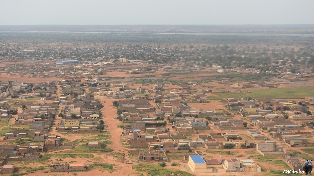 Das Bild zeigt die Luftaufnahme einer Stadt. In rechteckigen, eingemauerten Grundstücken stehen ein- und zweigeschossige graue Häuser mit Flachdach. Dazwischen verlaufen breite Straße aus rötlichem Sand. Zwischen den Häusern sind einzelne Grasflächen. Im Hintergrund stehen Baumkronen zwischen den Häusern und ein Flusstal verläuft parallel zum oberen Bildrand.