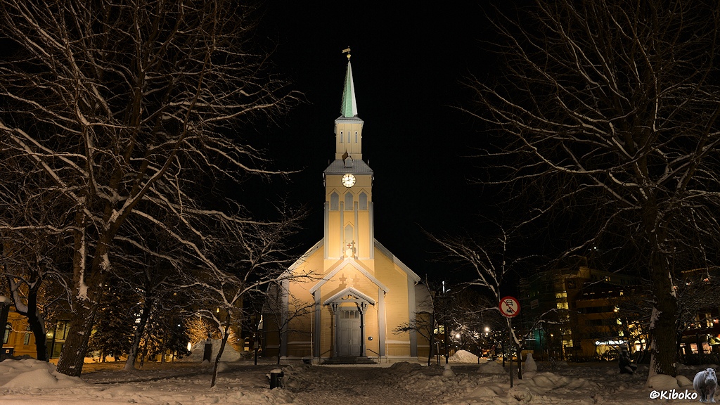 Das Bild zeigt ein gelbe Holzkirche mit schlanken Turm und hoher Turmspitze. Auf beiden Seiten stehen laublose Bäume mit Schnee auf den Ästen.