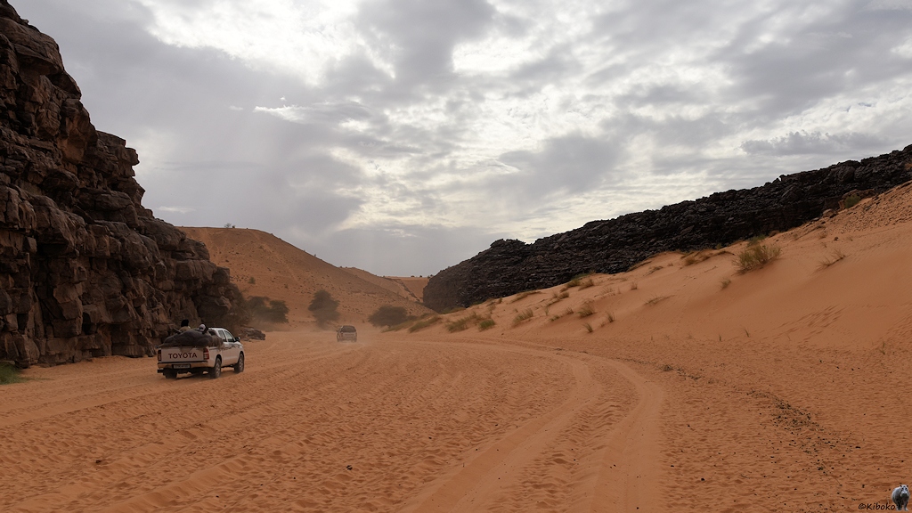 Das Bild zeigt zwei vorausfahrende Geländewagen im lachsfarbenen Tiefsand in einem engen Tal. Link ragt eine dunkle Felswand empor. rechts ist ein Hang aus lachsfarbenen Sand, der in eine schwarze Felswand übergeht. Am Himmel sind Wolken.