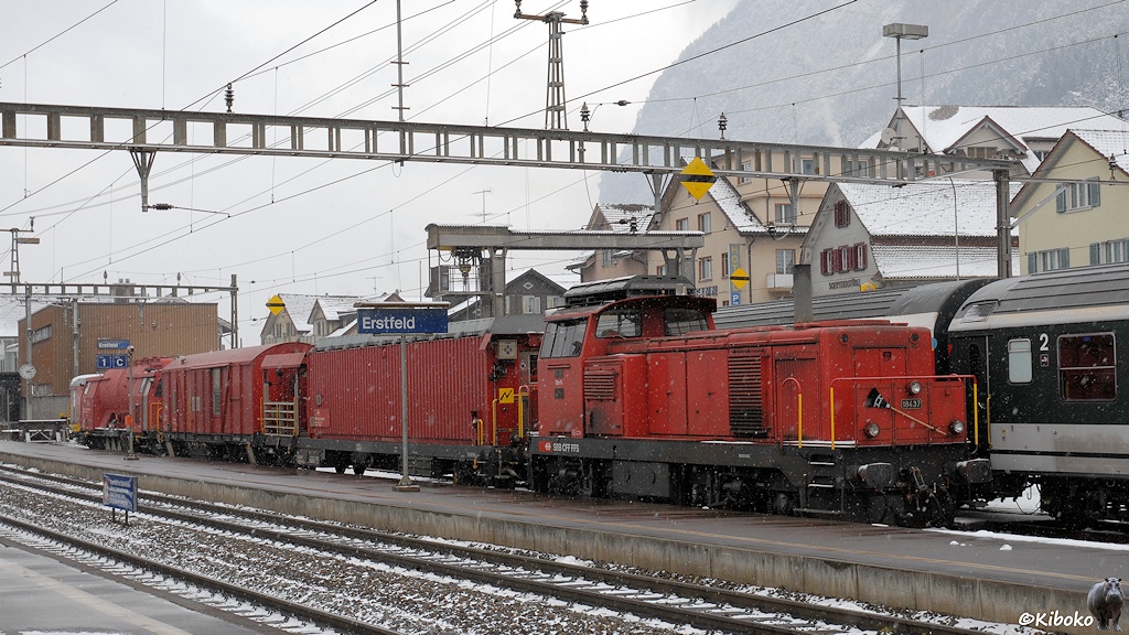 Das Bild zeigt eine rote vierachsige Diesellkomotive mit einen Hilfszug auf einem Abstellgleis im Bahnhof.
