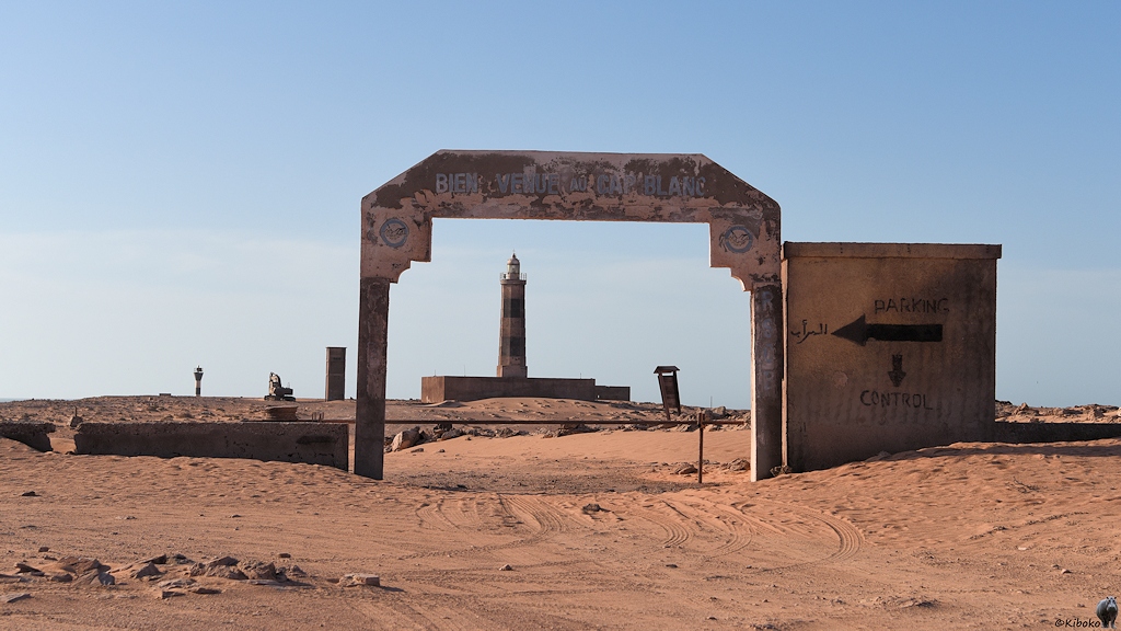 Das Bild zeigt ein eckiges Tor aus Beton mit abgeflachten Ecken im Gegenlicht. Durch das Tor führt ein Sandweg. Das Tor rahmt einen schwar-weiß geringelten Leuchtturm ein, der in großer Entfernung steht.