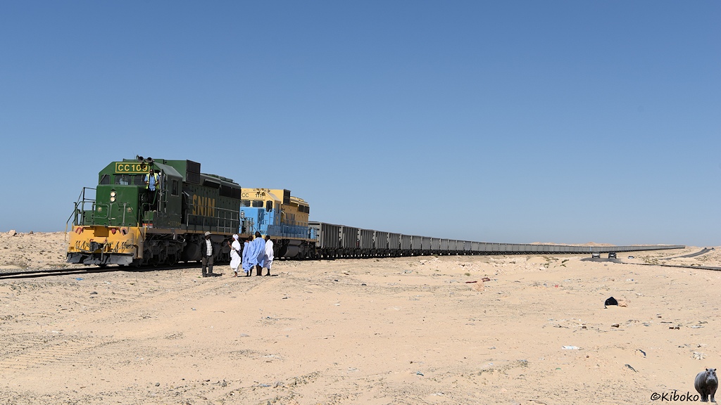 Das Bild zeigt eine grüne und eine beige-hellblaue Diesellok vor einer bis zum Horizont reichende Schlane von offenen Güterwagen. Vor der Ersten Lok steht eine Traube von Menschen in weißen und hellblauen Geändern. Daneben steht der Lokführer in einen schwarzen Outfit.