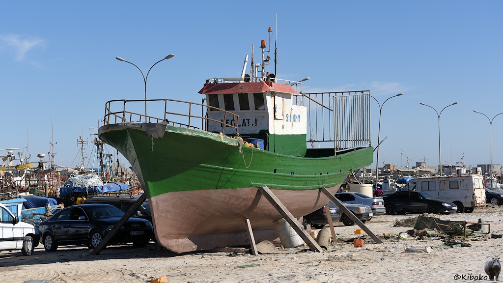 Das Bild zeigt ein auf dem Land aufrecht stehendes grünes Fischerboot mit einer Kajüte in Schiffsmitte. Holzbalken stützen das Boot. Die Aufbauen sind weiß. Der untere Teil des Rumpfes ins braun. Autos stehen neben dem Boot.