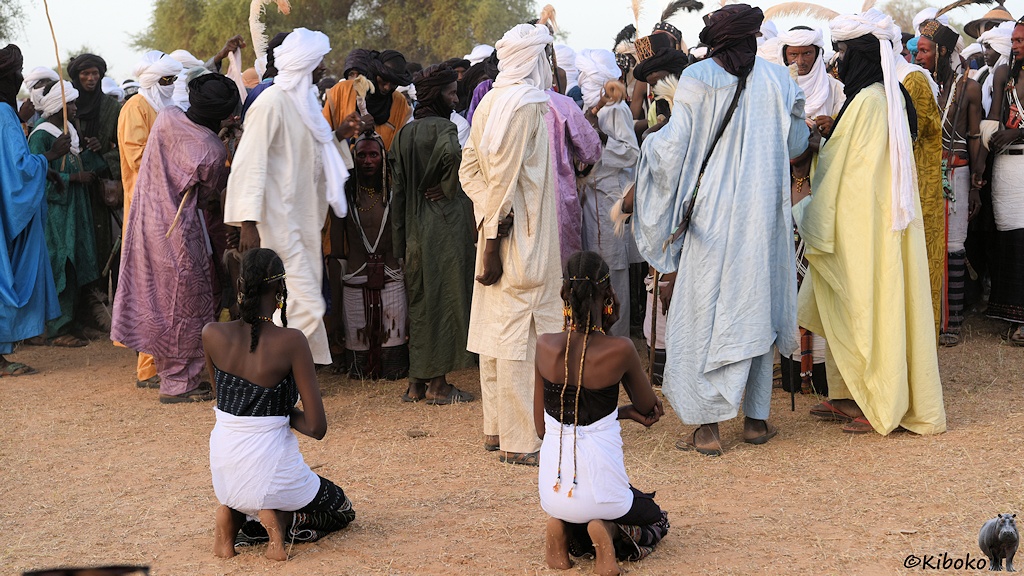 Das Bild zeigt zwei knieende junge Damen von hinten. Davor ist eine Gruppe von Männern in langen Gewändern. In der Gruppe sind die siegreichen Tänzer versteckt.