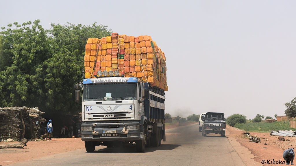 Das Bild zeigt einen weißen IVECO LKW. Auf der Ladefläche ist ein Berg mit gelben Kanistern, der das Führerhaus weit überragt. Der LKW bläst eine dunkelgraue Wolke aus dem Auspuff.