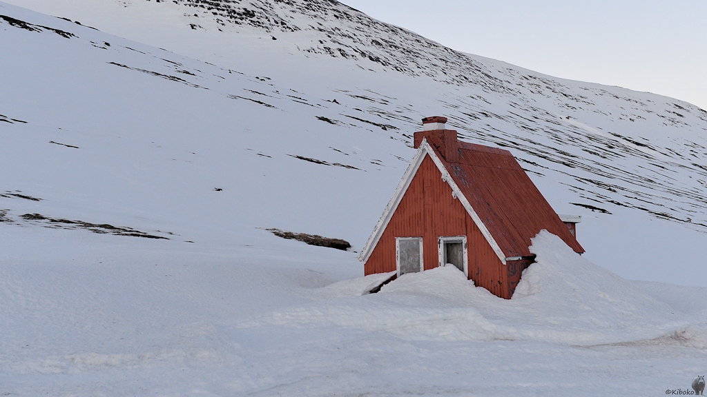 Ein kleines rotes Haus mit spitzen hohen Dach ist von hohem Schnee eingeschlossen.