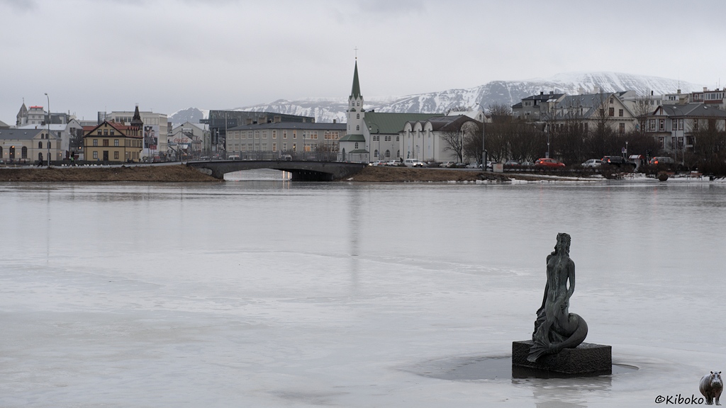 Ein See ist mit einer Eisfläche bedeckt. Im Vordergrund steht eine Statue im See. Im Hintergrund ist eine Straßenbrücke, Kirche, Häuser und schneebedeckte Berge unter einer Wolkendecke.