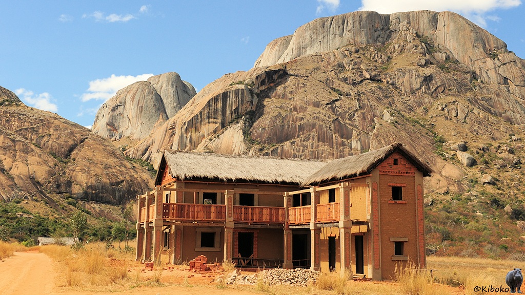 Das Bild zeigt ein zweigeschossiges, erdfarbenes Gebäude in Winkelform. Im ersten Stock ist ein breiter Balkon. Das Dach ist mit Stroh gedeckt. Hinter dem Haus ragen hohe Berge aus runden hellgrauen und braungrauen Fels empor.