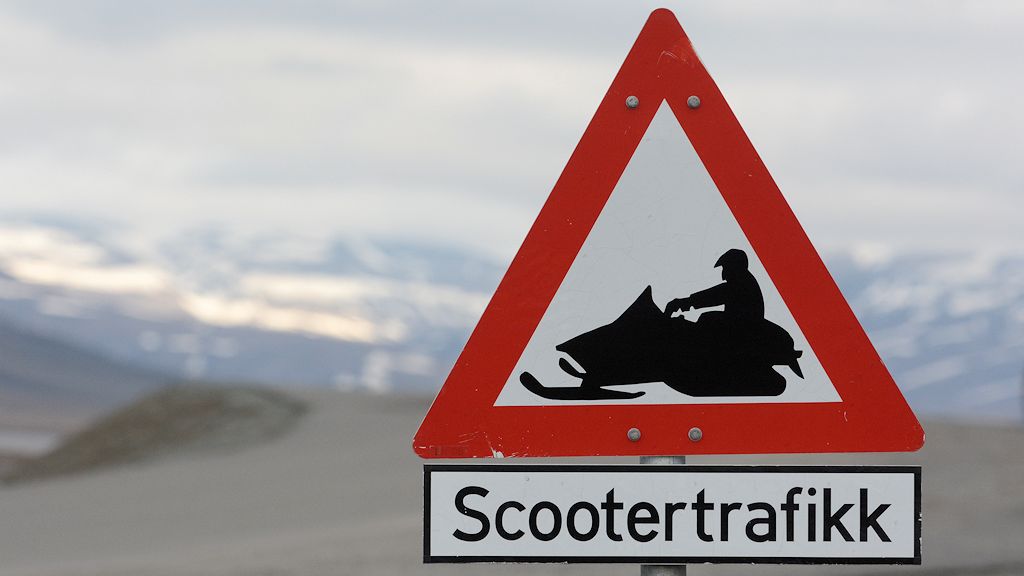 Das Bild zeigt ein dreieckiges Schild mit der Spitze nach oben und rotem Rand. Darauf ist ein schwarzes Schneemobil mit Fahrer auf weißem Grund. Darunter ist ein Zusatzschild mit dem Text: Scootertrafikk.