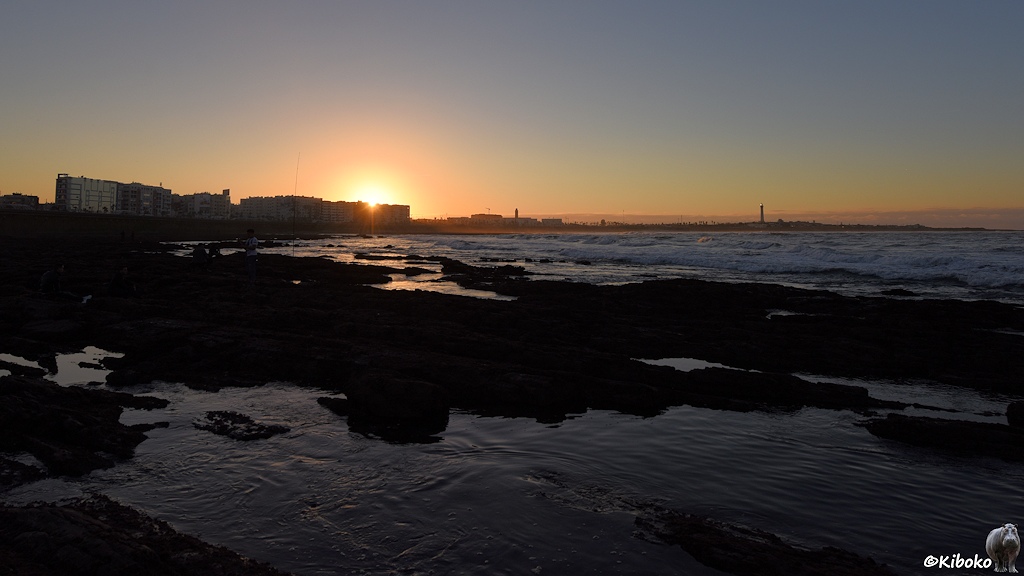 Das Bild zeigt eine Bucht bei Sonnenuntergang. Die Sonne geht hinter mehrgeschossigen Gebäuden unter. Am rechten Hintergrund steht ein Leuchtturm auf einer Halbinsel. Im Vordergrund sind Felsen mit Wasserflächen.