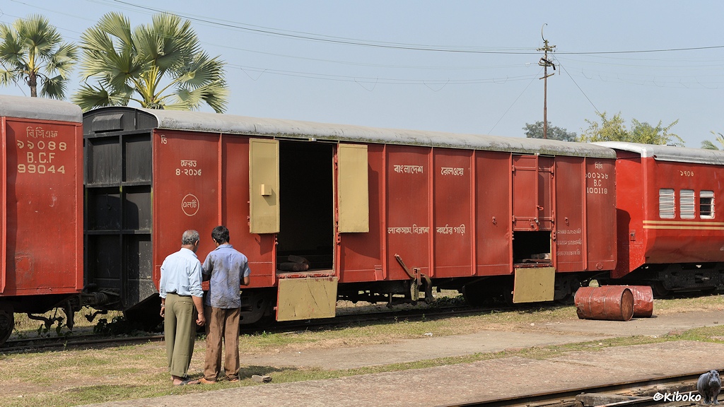 Das Bild zeigt einen braunen vierachsigen gedeckten Güterwagen Der Wagen hat schwarze Stirnseiten und ein silbernes Dach. Die linke Tür steht offen. Beide Flügel sind aufgekappt und das Unterteil ist nach unten geklappt. Bei der rechten Tür ist nur das Unerteil nach unten geklappt. Vor dem Wagen liegen zwei braune Fässer. Zwei Mann stehen mit dem Rücken zum Betrachter vor dem Wagen.